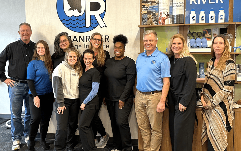 Staff and students pose together in front of the Urban River Massage Institute logo and product display shelves.